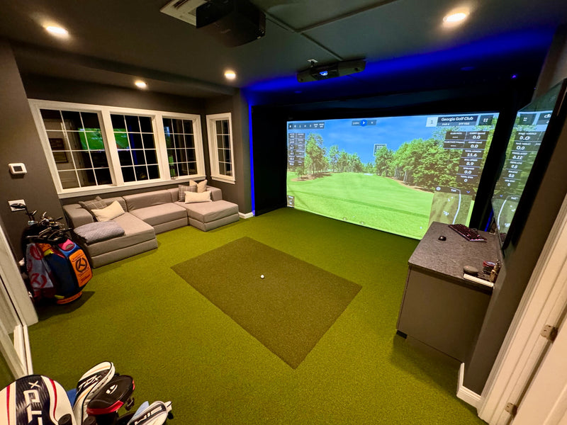 Indoor golf simulator room with large projection screen showing a virtual golf course, green turf floor, and seating area